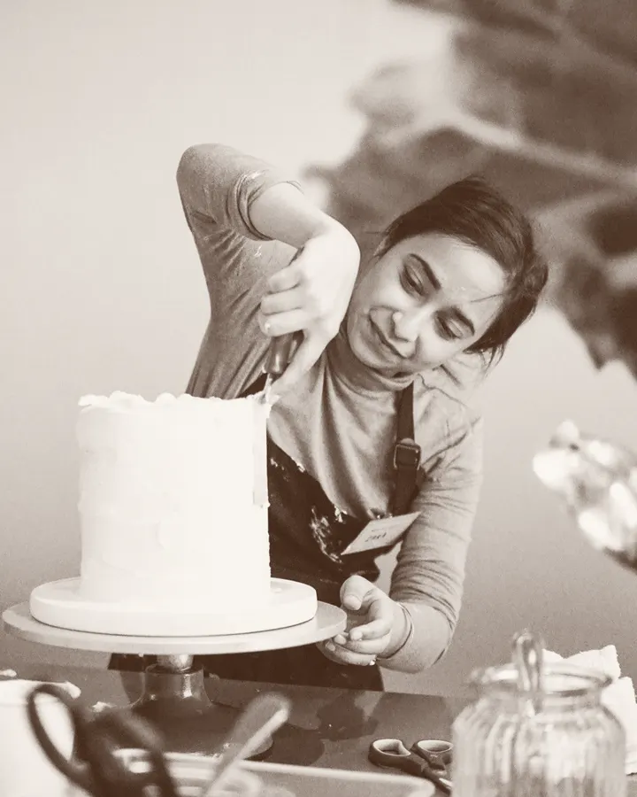 Dahlia leaning in to smooth buttercream on a tall white cake on a turntable.